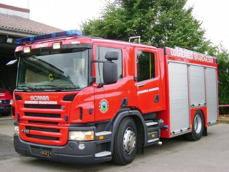 Rød Scania brandbil fra Odsherred Brandvæsen parkeret udenfor en bygning med grønne træer i baggrunden.