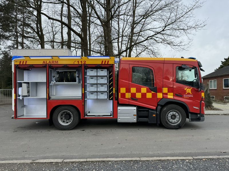 Rød brandbil med gule felter og åbne rum fyldt med brandudstyr holder parkeret ved en vej.