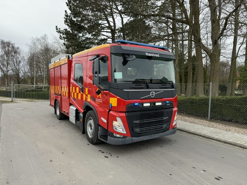 Rød brandbil fra Næstved Brand & Redning med gult stribemønster holder på en asfalteret vej foran træer.