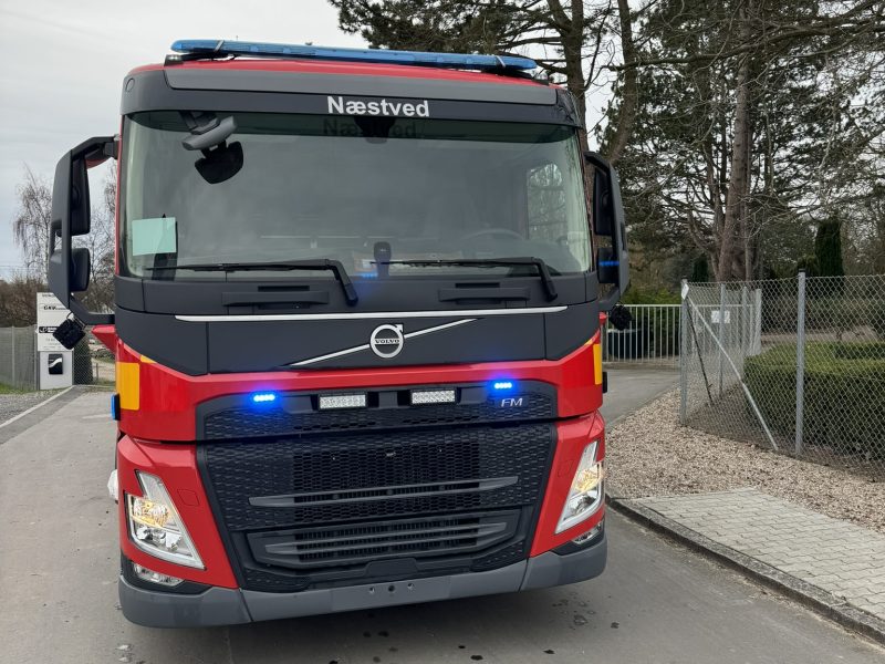 Fronten af en rød Volvo brandbil fra Næstved med blå udrykningslys tændt, parkeret på en grå asfaltvej.